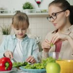 Cute loving daughter is helping mother with salad cutting cucumbers feeding young woman and laughing having fun at home. People and cookery concept.