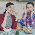 Joyful mother and cute cheerful daughter have fun grimacing silly with vegetables and looking into camera while cooking in the kitchen at home. Family, cook, and people concept